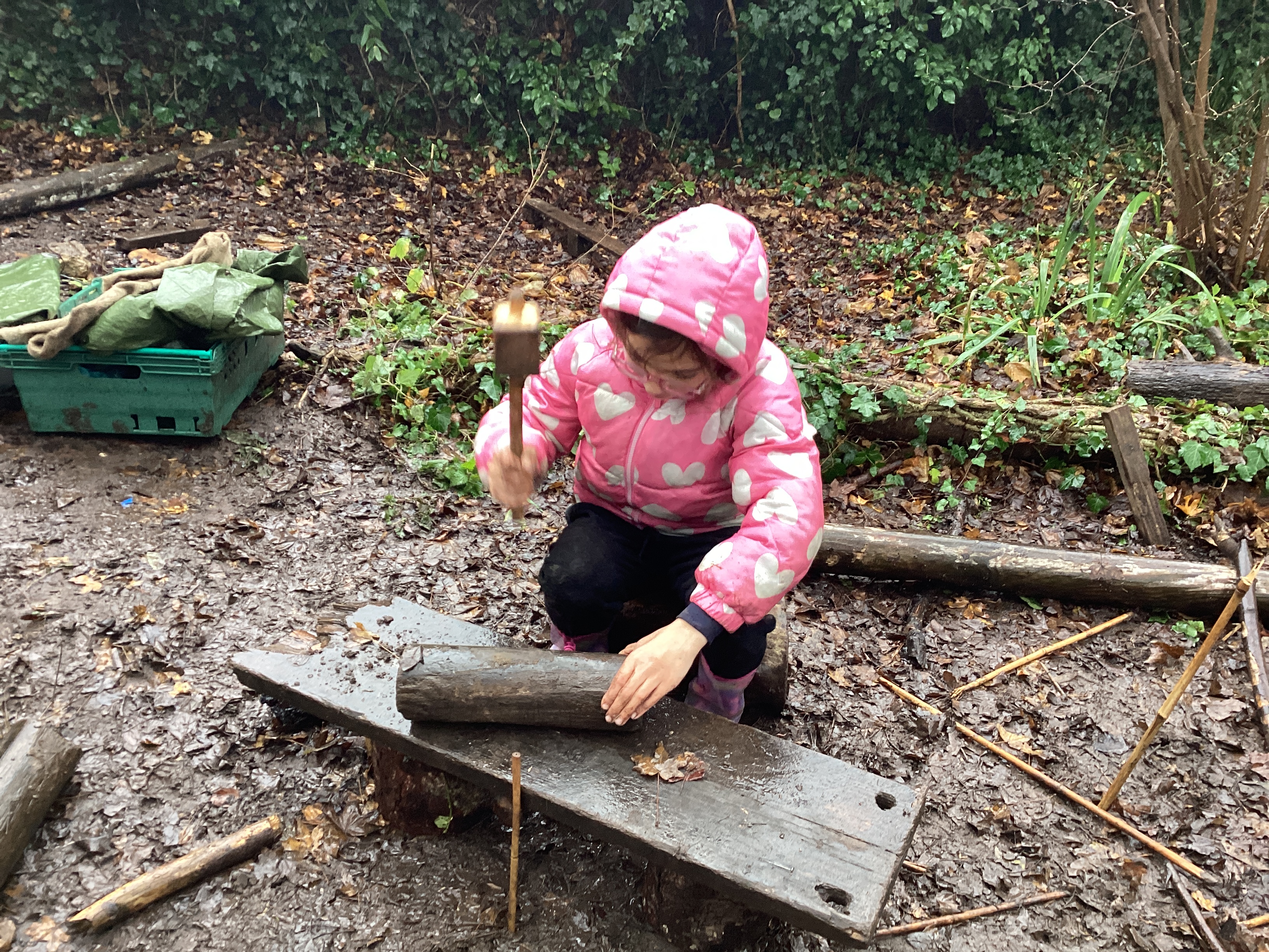 A child in a coat, using a hammer on a piece of wood.