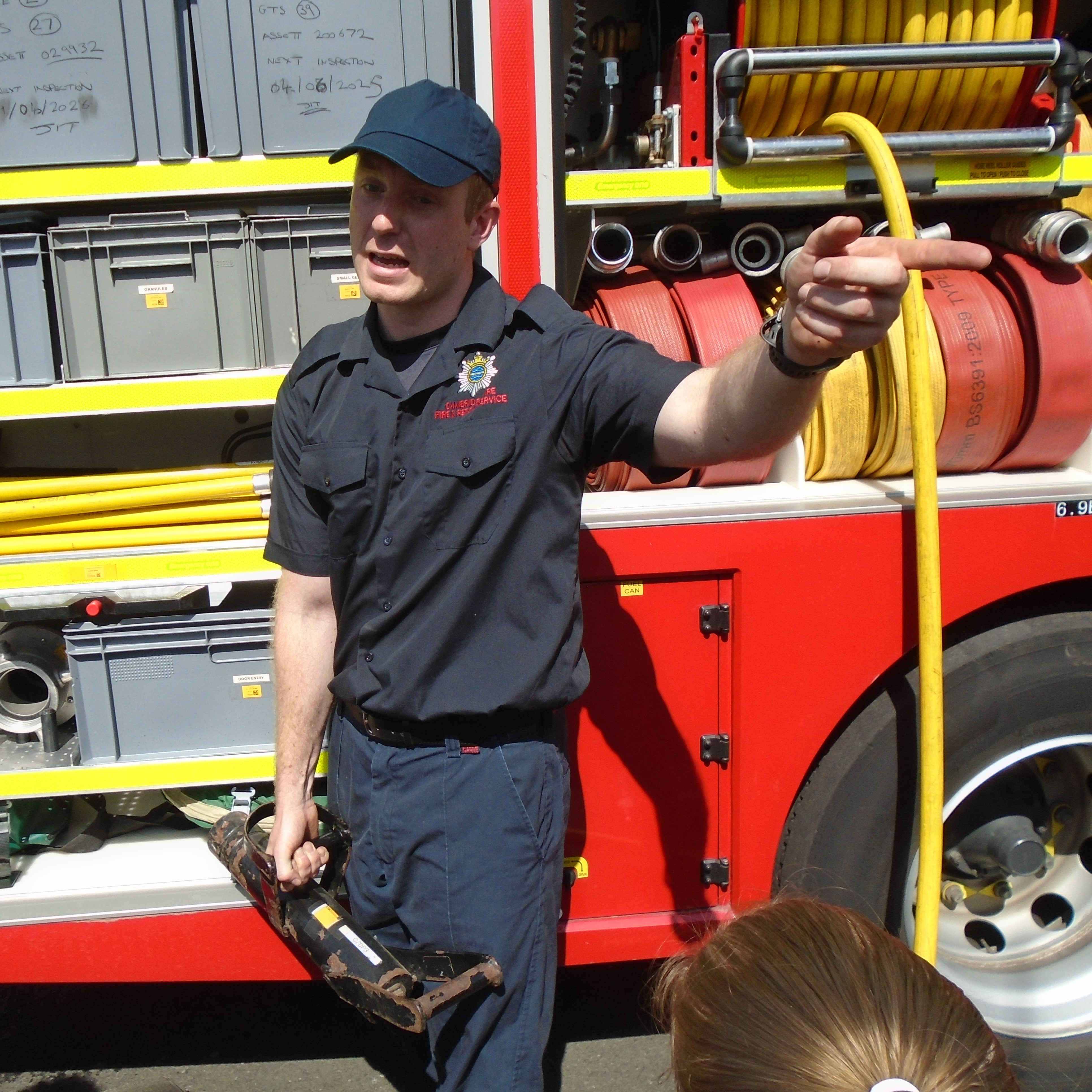 A fireman holding some equipment, talking to a group of children.